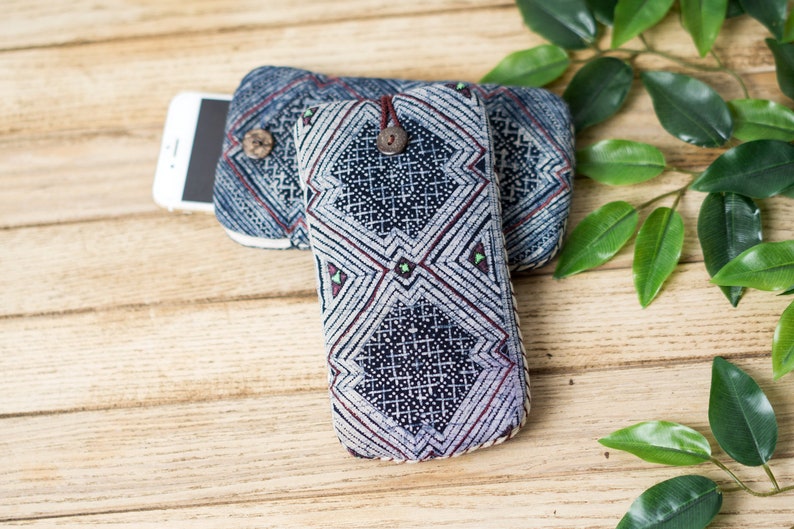 May include: Two blue and white patterned fabric phone cases with a wooden button closure. The cases are on a wooden surface with green leaves.