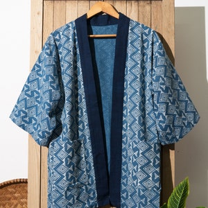 May include: A blue and white patterned kimono with a dark blue trim. The kimono is hanging on a wooden hanger against a wooden background.