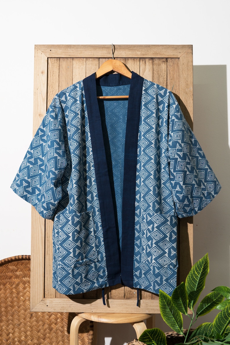 May include: A blue and white patterned kimono with a dark blue trim. The kimono is hanging on a wooden hanger against a wooden background.
