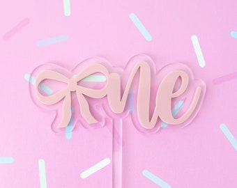 May include: Pink acrylic cake topper with the word "one" and a bow. The topper is on a pink background with white and blue sprinkles.
