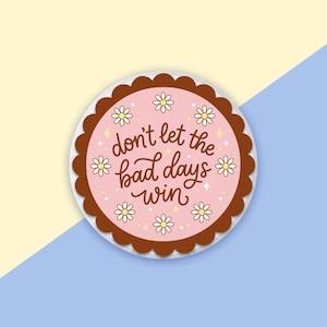 May include: A circular sticker with a pink centre and a brown, scalloped edge. The sticker displays the phrase "don't let the bad days win" in a handwritten style, surrounded by small daisy illustrations.