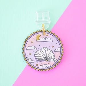 May include: A round, lilac-coloured earring charm featuring an illustration of an open book, clouds, stars, and a crescent moon. The charm has a gold-coloured scalloped edge and a clear earring post.