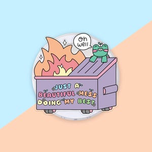 May include: A sticker featuring a purple dumpster on fire with a small green frog sitting on top. The text reads "OH WELL" in a speech bubble and "JUST A BEAUTIFUL MESS DOING MY BEST" on the side of the dumpster, decorated with small flowers.