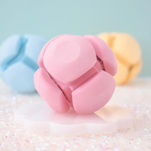 May include: A pink, spherical object with petal-like segments, on a white, cloud-shaped base. Two other similar objects, one blue and one yellow, are blurred in the background. The objects appear to be made of a smooth, matte material.