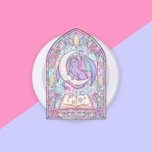May include: A sticker featuring a stained glass window design with a purple dragon perched on a crescent moon above an open book. The illustration includes pink roses, crystals, and stars in a pastel color palette with a holographic glitter effect.