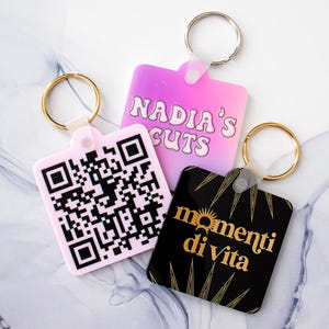 Peut inclure: Trois porte-clés avec des designs différents. Le premier porte-clés est rose avec un code QR blanc et noir. Le deuxième porte-clés est rose et violet avec le texte "Nadia's Cuts". Le troisième porte-clés est noir avec des accents dorés et le texte "momenti di vita".