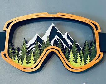 May include: Wooden ski goggles with a mountain scene inside. The goggles are painted in shades of brown, black, white, and green.
