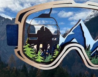 May include: A layered wooden wall art piece shaped like ski goggles. The frame is light wood, featuring a cutout silhouette of two people on a chairlift, dark blue mountain peaks, and green pine trees, creating a multi-dimensional scene.
