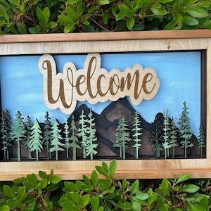 May include: A wooden framed wall art piece with a blue sky background, mountains, and a forest of green trees. The word "Welcome" is cut out of wood and placed above the mountains.