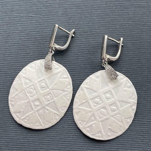 May include: A pair of white oval earrings with a geometric pattern. The earrings have silver-colored lever-back closures and small, rectangular accents with embedded clear stones. The earrings are displayed on a dark gray surface.