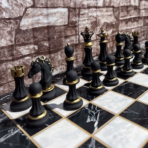 Könnte beinhalten: Ein schwarzes und goldenes Schachspiel auf einem schwarz-weiß karierten Brett. Die Schachfiguren sind schwarz mit goldenen Akzenten. Das Brett hat einen goldenen Rand. Der Hintergrund ist eine Backsteinmauer.