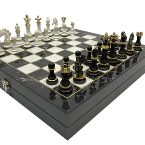 May include: A black and white marble chessboard with gold accents and black and white chess pieces. The board is closed and the pieces are set up for a game.