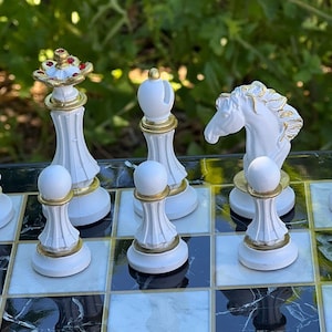 May include: White chess pieces on a black and white marble chessboard. The pieces are made of white ceramic with gold accents. The pieces include a king, queen, knight, rook, bishop, and pawn.