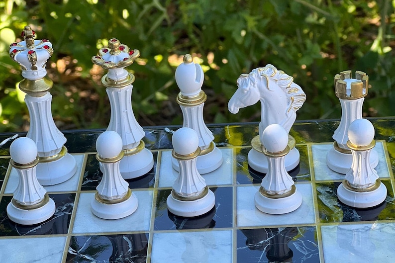 May include: White chess pieces on a black and white marble chessboard. The pieces are made of white ceramic with gold accents. The pieces include a king, queen, knight, rook, bishop, and pawn.