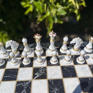 May include: A white chess set with gold accents on a black and white marble chessboard. The pieces are arranged in the starting position for a game of chess.