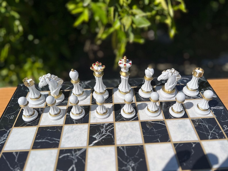 May include: A white chess set with gold accents on a black and white marble chessboard. The pieces are arranged in the starting position for a game of chess.