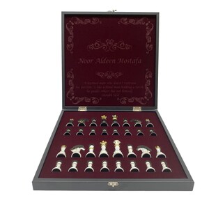 May include: A wooden chessboard set with a burgundy velvet interior and a personalized inscription that reads "Noor Aldeen Mostafa". The inscription is surrounded by a decorative border. A quote is also included: "A learned man who doesn't retain his position is like a bird that doesn't have a nest, he guides others but not himself."  The chess pieces are arranged on the board, ready for a game.
