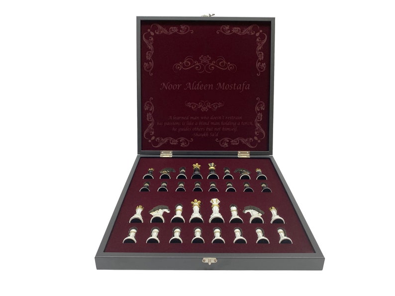 May include: A wooden chessboard set with a burgundy velvet interior and a personalized inscription that reads "Noor Aldeen Mostafa". The inscription is surrounded by a decorative border. A quote is also included: "A learned man who doesn't retain his position is like a bird that doesn't have a nest, he guides others but not himself."  The chess pieces are arranged on the board, ready for a game.