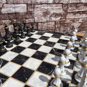 May include: A black and white marble chessboard with gold accents. The chess pieces are arranged in a starting position, with the white pieces on the right side of the board and the black pieces on the left side. The white pieces are made of white ceramic with gold accents, while the black pieces are made of black ceramic with gold accents.