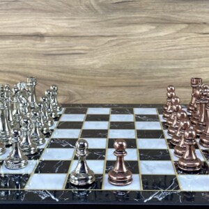 以下が含まれることがあります： 黒と白の大理石のチェス盤に、銀と銅のチェスの駒が置かれています。銀の駒は盤の左側、銅の駒は右側です。駒は標準の開始位置に配置されています。