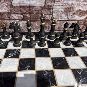 May include: A black and gold chess set with black and white marble board. The chess pieces are arranged in the starting position for a game of chess.
