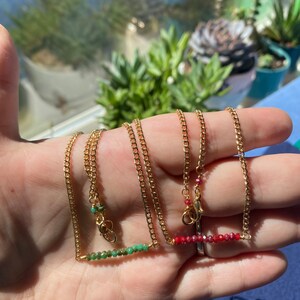May include: Two gold chain necklaces with a bar of green and red gemstones. The necklaces are on a hand.