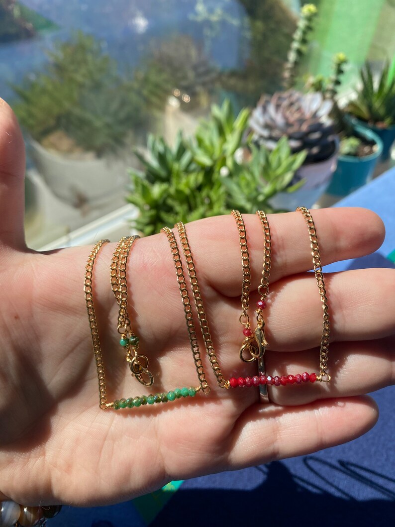 May include: Two gold chain necklaces with a bar of green and red gemstones. The necklaces are on a hand.