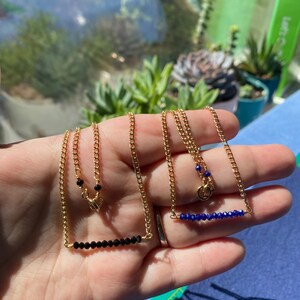 May include: Two gold chain necklaces with a bar of gemstones. One necklace has black gemstones and the other has blue gemstones.