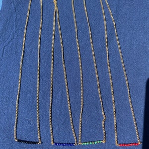 May include: Four gold chain necklaces with different colored gemstone bars. The necklaces are laid out on a blue fabric surface. The gemstones are black, blue, green, and red.