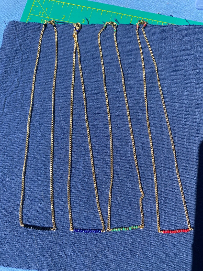 May include: Four gold chain necklaces with different colored gemstone bars. The necklaces are laid out on a blue fabric surface. The gemstones are black, blue, green, and red.