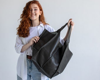 Puede incluir: Una mujer con el pelo largo y rizado rojo sonríe y sostiene una gran bolsa de mano de cuero gris oscuro.