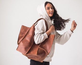 Puede incluir: Una mujer con una sudadera con capucha beige claro lleva un gran bolso de mano de cuero marrón.