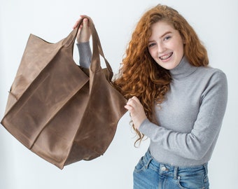 Puede incluir: Una mujer con el pelo largo y rizado rojo está sosteniendo una gran bolsa de mano de cuero marrón. Está sonriendo y lleva un jersey de cuello alto gris y unos vaqueros azules.