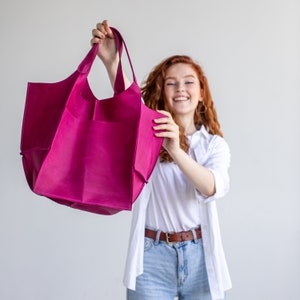 May include: A woman holds a large, bright pink leather tote bag with a wide opening and multiple pockets. The bag is made of soft, supple leather and has a casual, relaxed style.