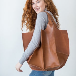 May include: A woman with long, curly red hair is carrying a large, brown leather tote bag. The bag has a wide, open top and a structured, boxy shape.