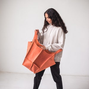May include: A woman is holding a large, orange leather tote bag with a single handle. The bag is folded in on itself, and the woman is looking at it.