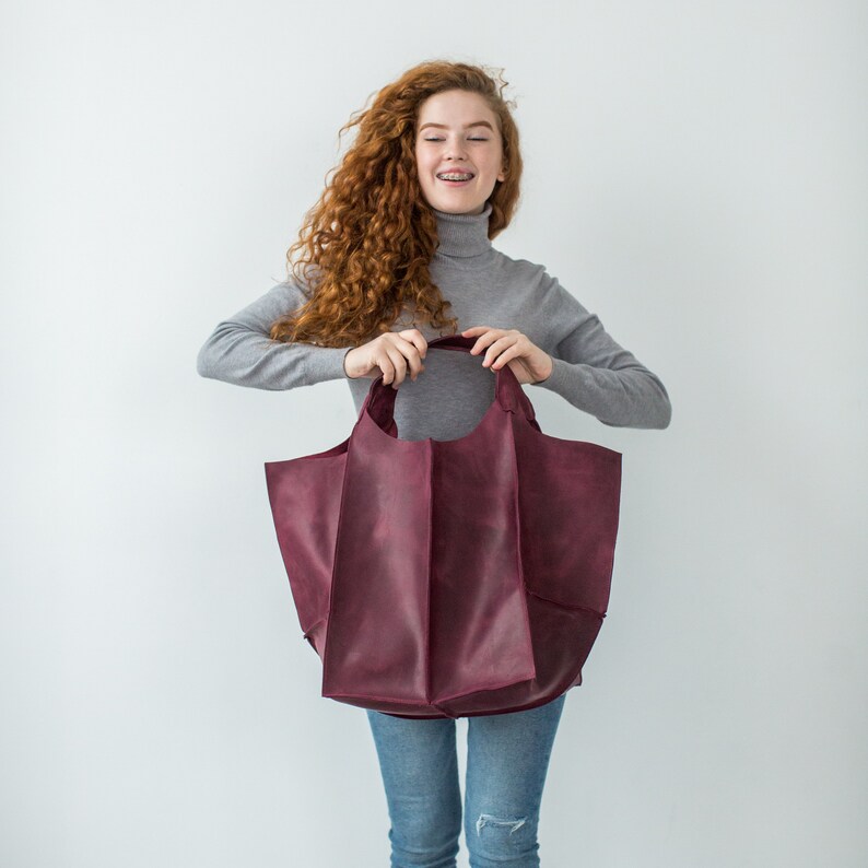 May include: A woman with long, curly red hair is holding a large, burgundy leather tote bag. The bag has a wide, open top and a structured, boxy shape.