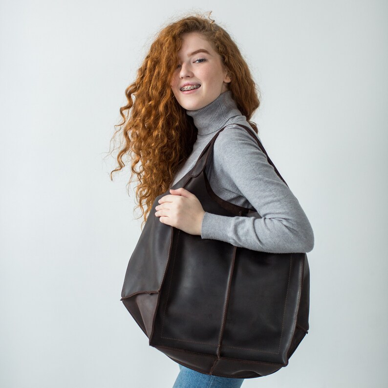 May include: A woman with long, curly red hair is wearing a gray turtleneck sweater and carrying a large brown leather tote bag.