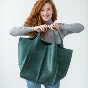 May include: A large, green leather tote bag with a wide opening and a structured shape. The bag has a single handle and is held by a person.