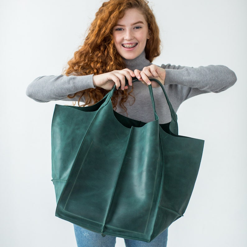 May include: A large, green leather tote bag with a wide opening and a structured shape. The bag has a single handle and is held by a person.