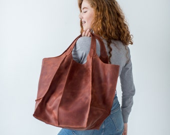 Può includere: Una donna con lunghi capelli rossi ricci porta una grande borsa tote in pelle marrone sulla spalla. La borsa ha una parte superiore larga e aperta e un design semplice.