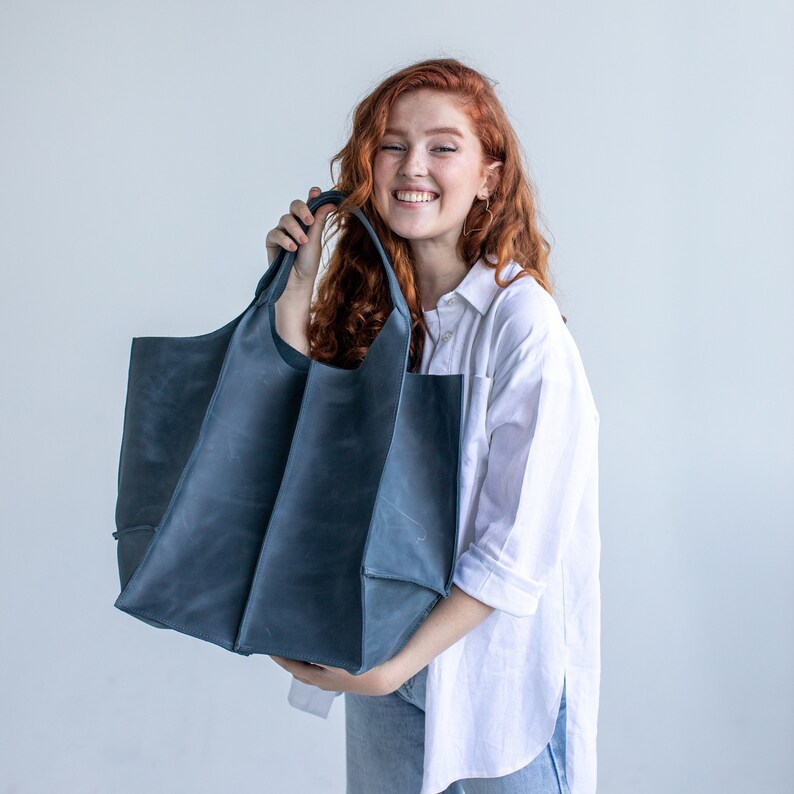 May include: A woman is holding a large, dark blue tote bag. The bag has a wide opening and a structured shape. The bag is made of a durable material and has a simple design.