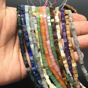 May include: A strand of 12 different colored gemstone beads, each bead is a square shape. The beads are strung on a clear thread. The colors of the beads are green, blue, purple, brown, orange, pink, and white.