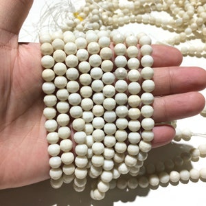 Puede incluir: Primer plano de cuentas facetadas de color crema ensartadas en un hilo, sostenidas en una mano. Las cuentas tienen un acabado mate y un tamaño uniforme. Se ven más hilos de cuentas en el fondo.