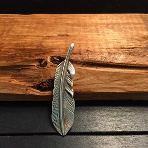 May include: A silver-colored feather pendant with detailed veining. The pendant is displayed against a textured, brown wooden background. The feather design is a popular motif in jewelry and accessories.