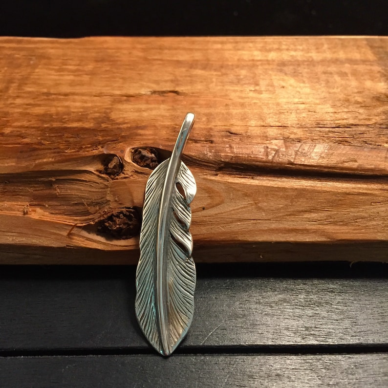 May include: A silver-colored feather pendant with detailed veining. The pendant is displayed against a textured, brown wooden background. The feather design is a popular motif in jewelry and accessories.