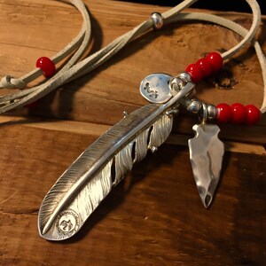 May include: A silver feather pendant necklace with a small silver arrowhead charm and red beads. The necklace is on a light brown leather cord.