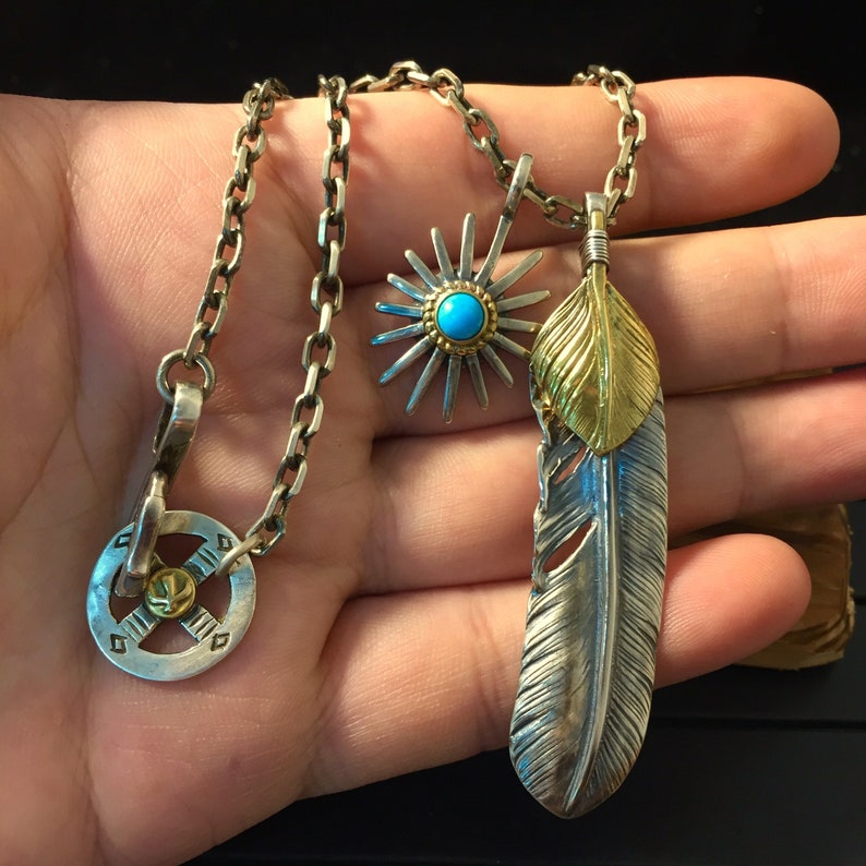 May include: A silver chain necklace with three pendants: a silver starburst with a turquoise stone, a silver and gold feather, and a silver circular pendant with a cross design.