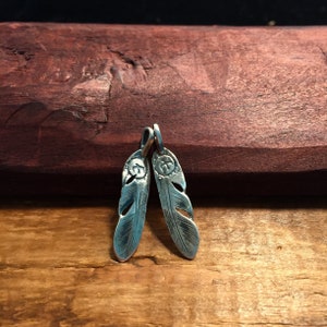 Puede incluir: Dos encantos de plumas de plata con un dise&ntilde;o detallado. Las plumas cuelgan de una superficie de madera.
