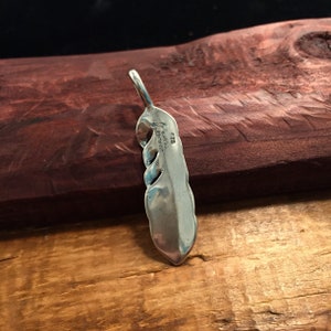May include: A sterling silver pendant in a feather design. The pendant has a loop at the top for a chain and a curved, openwork design. The back of the pendant is stamped with "STERLING" and "925". The pendant is displayed on a wooden surface.